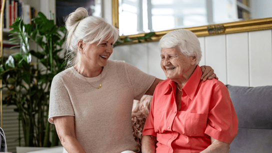 Photo of two people, with white hair, sitting on couch, smiling at each other